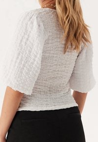 Haut blanc texturé avec dos smocké et larges manches bouffantes. Présente une taille ajustée et un tissu doux froissé. Associé à un pantalon foncé à rayures.