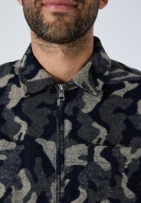 Een bebaarde man in een ritssluiting jack met een donkerblauw en grijs camouflagepatroon en een kraag, tegen een effen lichte achtergrond.
