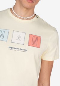 Camiseta de algodón amarillo claro con gráficos cuadrados en azul, blanco y coral con el texto: "¡Peligro! Zona de tiburón Harper: Cuida tus aletas y amigos."