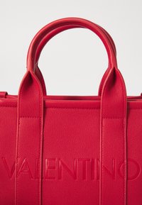 Bolsa de mão de couro vermelho com acabamento texturizado, apresentando o logotipo "VALENTINO" em relevo e duas alças robustas e arredondadas.