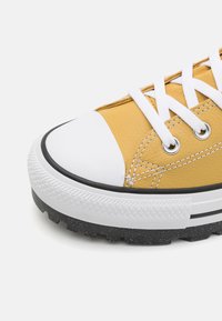 Žluté tenisky s texturovaným koženým svrškem, bílým gumovým špičkovým krytem, výraznou černou podrážkou a bílými tkaničkami. Mají kontrastní prošívání.