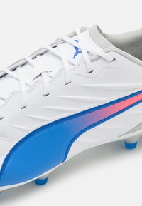 Scarpe da calcio bianche con accenti blu e rosa, dotate di una tomaia sintetica testurizzata, una linguetta grigia e un design tradizionale con lacci.