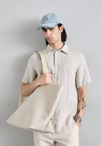 Lichtbeige linnen overhemd met korte mouwen, houdt een grote beige totebag met een stevige band vast en draagt een lichtblauwe denim pet met een rafelige boord.