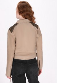 Chaqueta de cuero beige con acentos de encaje negro en los hombros, con un diseño ajustado y puños con cremallera. Textura suave, estilo corto.