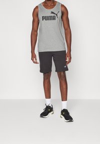 Grijze tanktop met vet zwart "PUMA" logo, gecombineerd met zwarte shorts met een klein logo. Model draagt zwarte sportschoenen en witte sokken.