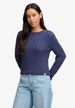 Femme aux longs cheveux foncés portant un pull en maille torsadée bleu marine et un jean bleu clair, debout avec les mains derrière le dos.
