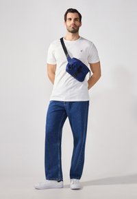 Blå crossover taske med sort rem, tekstureret stof, lynlås lukning og accent detaljer. Modellen bærer en hvid t-shirt og blå jeans.