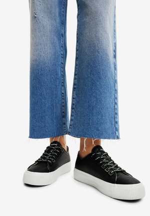 Sneakers neri con tomaia in pelle testurizzata, suola in gomma bianca a contrasto e lacci larghi e piatti, indossati con jeans a gamba larga blue con orlo sfrangiato.