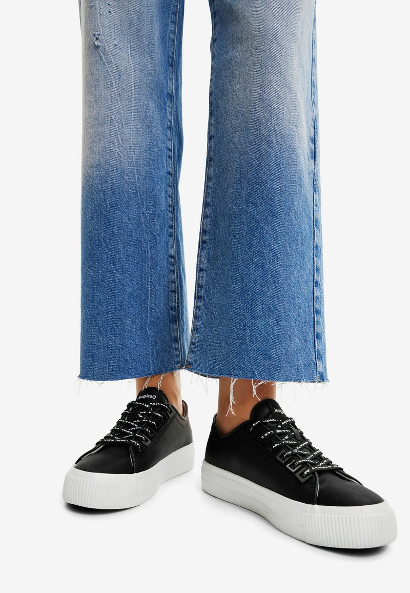 Sneakers neri con tomaia in pelle testurizzata, suola in gomma bianca a contrasto e lacci larghi e piatti, indossati con jeans a gamba larga blue con orlo sfrangiato.