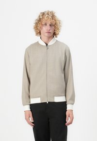 Beige bomberjakke med tekstureret stof, lynlåslukning, hvid krave og manchetter samt to hvide detaljeringer i kanten.