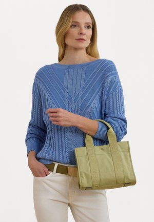 Femme portant un pull en maille torsadée bleu et un pantalon blanc, tenant un sac cabas vert clair avec deux anses et détail du logo.