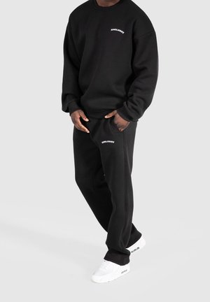 Man wearing black Smilodox sweatshirt and matching sweatpants with white sneakers, standing with one hand in pocket.