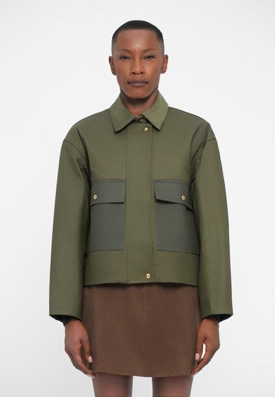 Chaqueta acortada color verde oliva con dos bolsillos frontales, accesorios dorados, cuello de camisa y textura suave. Lleva puesta sobre una falda marrón.