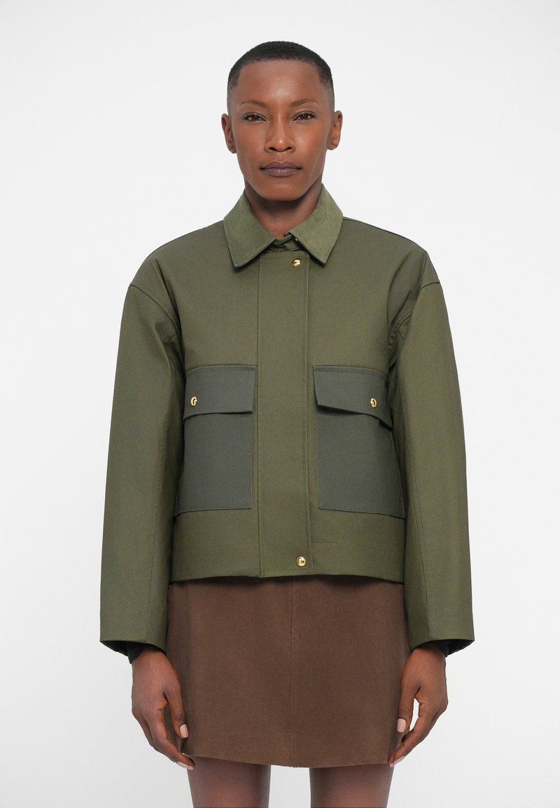Olive green cropped jacket with two front pockets, gold hardware, a shirt collar, and a smooth texture. Worn over a brown skirt.
