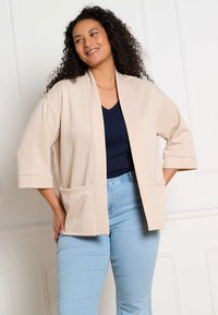 Frau mit lockigem Haar lächelt, trägt eine beige offene Strickjacke, ein dunkelblaues Oberteil, hellblaue Jeans und eine goldene Kettenhalskette, steht vor einer weißen Wand.