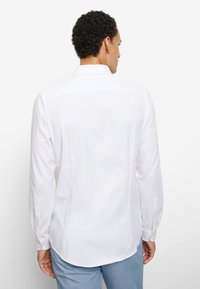 Homme portant une chemise blanche à manches longues et un pantalon bleu clair, vu de dos sur un fond blanc uni.