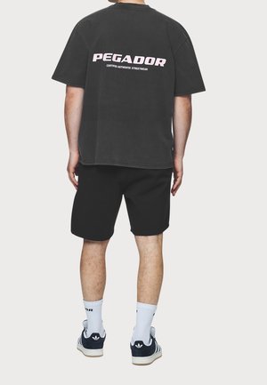 Persona de pie de espaldas, con una camiseta negra "PEGADOR", pantalones cortos negros, calcetines blancos y zapatillas negras Adidas.