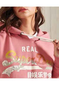 Rosa hoodie med framsidan i grafisk design, som har texten "SuperDry" i flera färger. Inkluderar dragsnören och ribbade ärmslut. Mjuk textur.