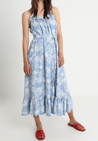 Niebieska długa sukienka w kwiaty z falbaniastym dekoltem, regulowanymi ramiączkami, marszczonym pasem i warstwowym dołem. Połączona z czerwonymi sandałami wsuwanymi.