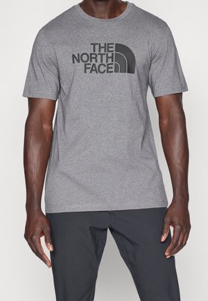 Homme portant un t-shirt gris à manches courtes avec un logo noir "THE NORTH FACE" et un pantalon noir, debout devant un fond blanc uni.