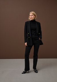 Šviesiaplaukė moteris stovi, dėvi juodą dvieilių sagų švarką, juodą golfą, juodus kelnes, pilkas kojines ir juodus aukštakulnius, stovėdama prieš rudą foną.