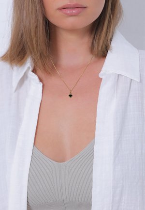 Guldhalskæde med en grøn ædelsten, båret over en lys grå ribbet top, lagd under en hvid linnedskjorte. Klar, minimalistisk design.