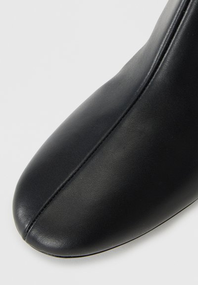 Bottine en cuir noir à bout pointu, texture lisse et un subtil détail de couture sur le devant. Design classique et minimaliste.