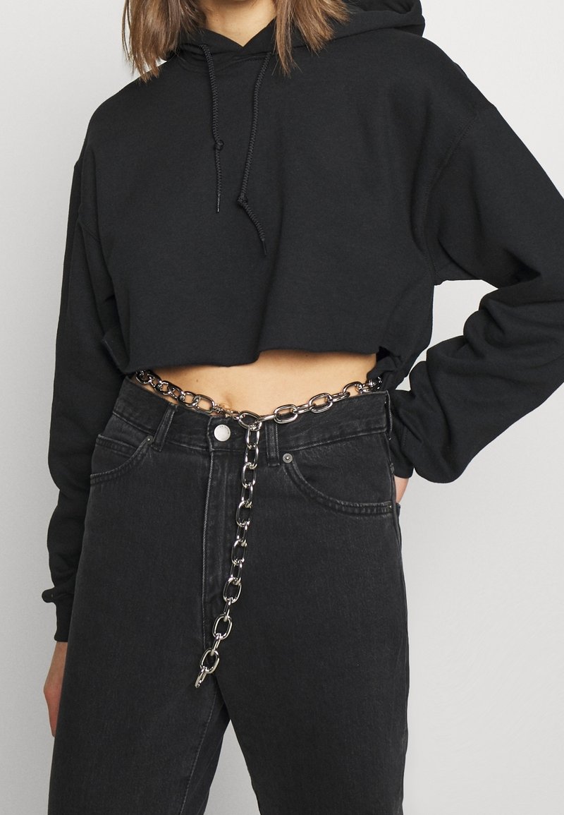 Donna che indossa un hoodie corto nero, jeans scuri a vita alta e una cintura a catena argentata con la parte finale pendente.