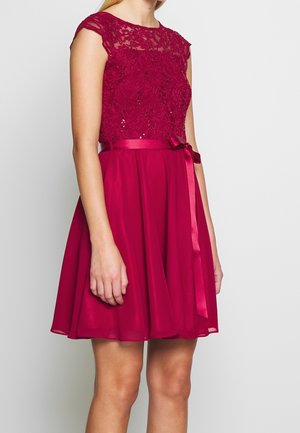 Frau trägt ein ärmelloses rotes Kleid mit Spitze und Pailletten auf dem Oberteil und einem Satinband, das an der Taille gebunden ist.