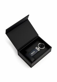 Metalinis raktų pakabukas su apvaliu žiedu ir tamsiai mėlynu odiniu dirželiu, ant kurio užrašyta "BOSS". Pateikta elegantiškoje juodoje dėžutėje.