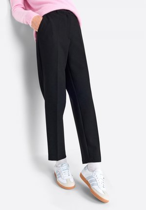 Person iført sorte bukser med lige ben, hvide sneakers med gummisåler, hvide sokker og en pink sweater med hånden i lommen.