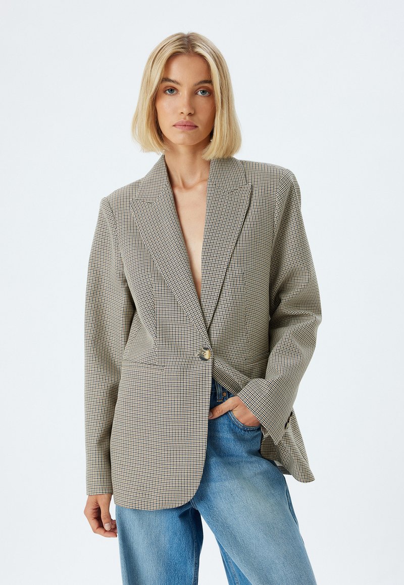 Geverifieerde blazer in lichtbruin en zwart, met een enkele knoopsluiting, notch-revers, gestructureerde schouders, gecombineerd met lichtblauwe jeans.