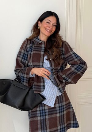 Femme souriante, portant une veste et une jupe à carreaux marron et bleus, tenant un sac à main noir et des lunettes de soleil, debout contre un mur blanc.