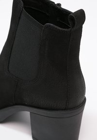 Gros plan sur une botte noire en cuir pour cheville avec panneau latéral élastique et talon bloc épais sur fond blanc.