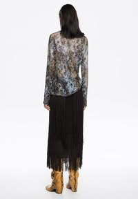 Długi rękaw, wzorzysty top w niebiesko-brązowych tonacjach, zestawiony z czarną spódnicą z frędzlami i żółtymi botkami z motywem wężowym, widziany od tyłu.