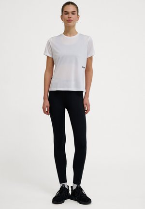 Hvid kortærmet t-shirt med rund hals og lille sort logo, parret med højtaljede sorte leggings og sorte atletiske sko.