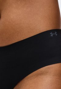 Κοντινό πλάνο της μέσης ενός ατόμου που φοράει μαύρα εσώρουχα Under Armour, εμφανίζοντας την υφή του δέρματος και λεπτομέρειες του λογότυπου στη ζώνη.