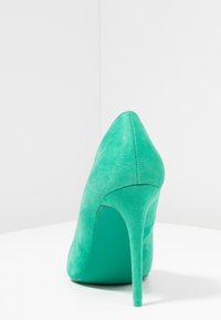 Escarpin en daim vert avec un bout pointu et un talon slim et haut. L'intérieur a un accent teal contrastant. Texture lisse.