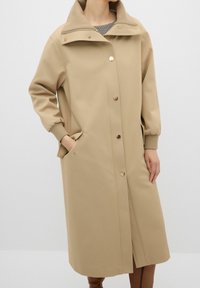 Abrigo beige a la altura de la rodilla, fabricado en un tejido suave, con un cuello alto, cierres de botones dorados y puños elásticos con bolsillos laterales.