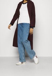 Person in einem langen dunkelroten Mantel, weißem Pullover mit schwarzen Bündchen, blauen Jeans und weißen Sneakers mit roten Akzenten, steht vor einem schlichten Hintergrund.