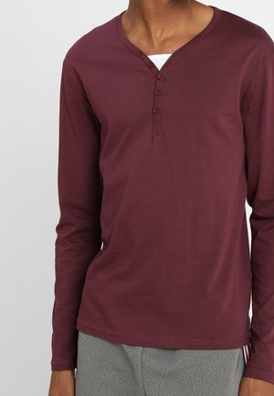 Mężczyzna ubrany w długi rękaw bordowej koszulę henley z czterema guzikami, na białej podkoszulce i szarych spodniach.