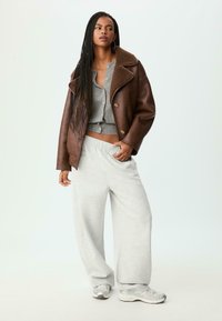 Giovane donna con lunghe trecce, che indossa un giubbotto di shearling marrone, un cardigan grigio con bottoni, pantaloni della tuta leggeri grigio chiaro e sneakers argentee.