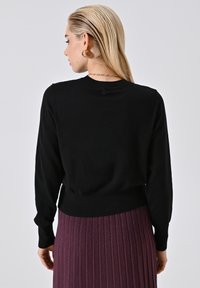 Blonde Frau, die einen schwarzen Langarm-Pullover und einen lila gerippten Rock trägt und mit dem Rücken zu einem schlichten hellgrauen Hintergrund steht.