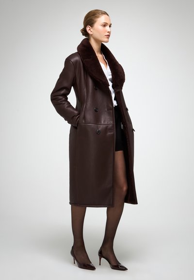 Palton din piele maro, cu două rânduri de nasturi și un guler mare din blană închisă la culoare, prezintă buzunare laterale și linii elegante, atingând lungimea de mijlocul gambei.