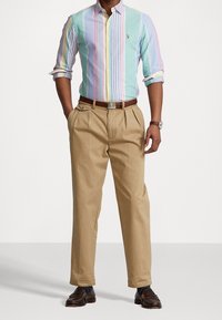 Camisa de rayas multicolores para hombre en azul claro, rosa y verde, con cuello y mangas enrolladas, combinada con pantalones plisados en color beige.