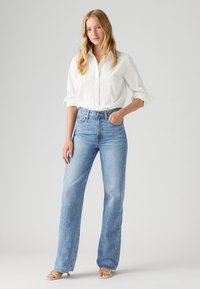 Jean taille haute bleu clair avec une coupe décontractée, associée à un chemisier blanc avec des détails brodés, et des sandales à lanières.