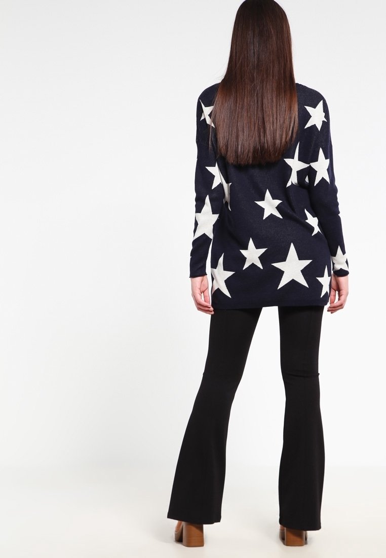Granatowy sweter z długim rękawem w duże białe gwiazdy, zestawiony z czarnymi spodniami dzwonami. Gładka faktura, dopasowany krój i dłuższa długość.