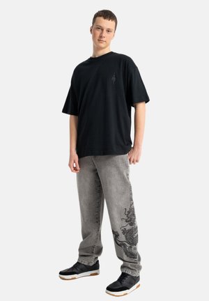 Młody mężczyzna stoi ubrany w luźny czarny t-shirt, szare dżinsy z czarnym wzorem smoka na jednej nodze oraz czarne trampki z białymi podeszwami.