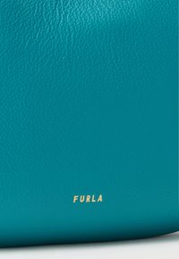 Surface en cuir texturé turquoise avec un petit logo doré "FURLA" centré près du bord inférieur.