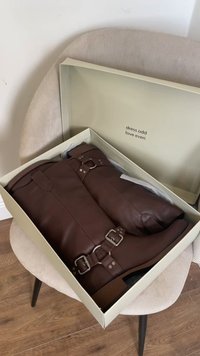 Botas de couro castanho com fivelas prateadas, dentro de uma caixa verde clara. A caixa tem um interior creme e uma frase impressa na tampa.